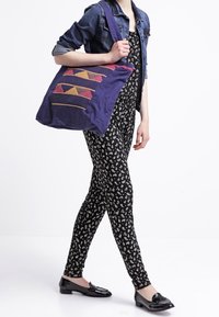 Bolsa de tela púrpura con acentos textiles geométricos en rosa y amarillo. Presenta una correa ancha y una textura suave.