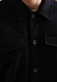 Sort knap-op jakke med krave, der har to forlommer og runde knapper. Lavet af tekstureret stof, minimalistisk design.