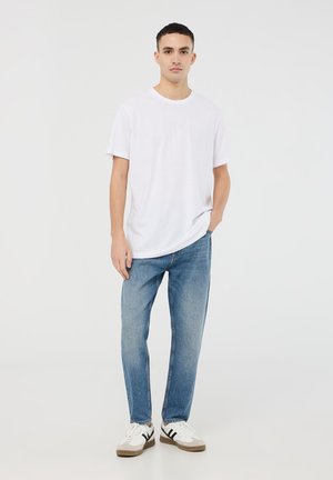 T-shirt bianco a maniche corte, vestibilità comoda, abbinato a jeans di denim blu chiaro e sneakers bianche con dettagli marroni, in piedi su uno sfondo neutro.