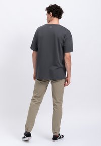 Grijze t-shirt met korte mouwen en ronde hals, voorzien van een klein logo op de achterkant, gecombineerd met beige broek en zwarte sneakers. Gladde stof textuur.