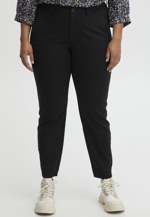 Černé tailored kalhoty mají slim fit, hladkou texturu a zapínání na knoflík. Nosí se s bílými teniskami, což ukazuje na ležérní vzhled.