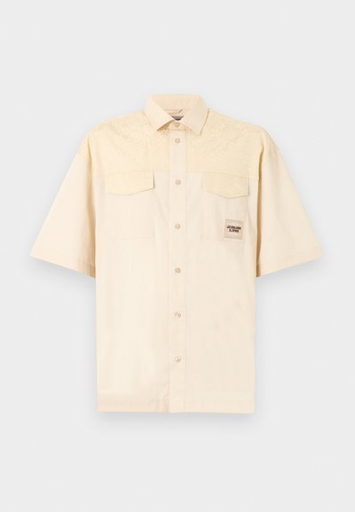 Chemise beige à manches courtes avec col pointu, deux poches à rabat sur le devant et une petite écusson rectangulaire sur la poitrine gauche.