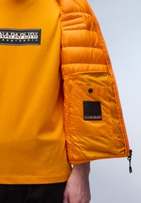 Żółta koszulka z czarnym logo, w połączeniu z pomarańczową kurtką puchową z kieszenią i zamkami błyskawicznymi na mankietach.