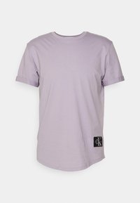 Fialové tričko s krátkým rukávem vyrobené z měkké bavlny. Obsahuje ohrnuté manžety a černou nášivku s logem a bílým textem v dolním levém rohu.