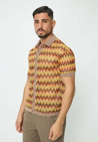 Polo à manches courtes avec un motif en zigzag orange, jaune et marron, avec un col beige et des boutons. Texture douce.