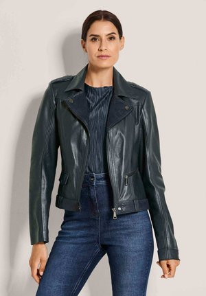 Frau mit dunklem Haar, die eine schwarze Lederjacke, ein strukturiertes schwarzes Top und dunkle blaue Jeans trägt und vor einem einfarbigen beigen Hintergrund steht.