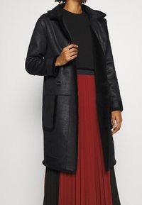 Svart shearlingjacka med krage, knäppning och två framfickor. Har en mjuk textur och en skräddarsydd passform som sträcker sig till mitten av låret.