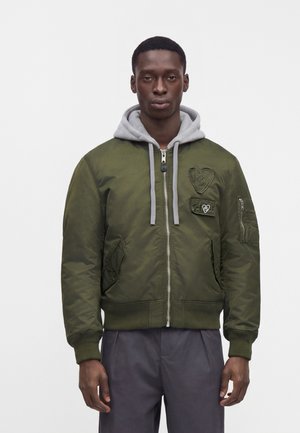 Un homme porte une veste bomber vert olive à fermeture éclair, avec une capuche grise, des écussons sur la poitrine et une poche sur la manche, sur fond uni.