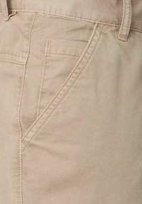 Beige katoenen broek met een diagonaal zakontwerp en zichtbare stikdetails. De stof lijkt glad met een lichte glans.