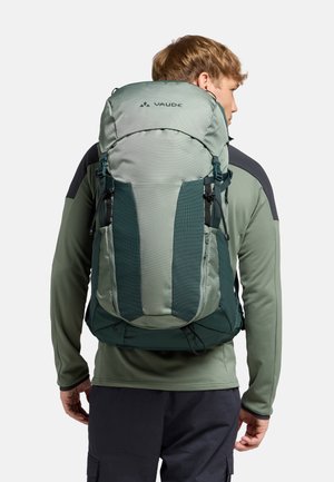 Zaino in tessuto verde con spalline regolabili, dettagli in rete e chiusura arrotolabile. Presenta diverse tasche con zip per lo stoccaggio.
