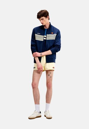 Jonge man met een marineblauwe jas met rits en lichtgele korte broek, staand met gekruiste armen, met kleine tatoeages op zijn linker dij.