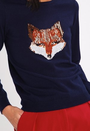 Marineblå sweater med et glasagtigt rævehoved-design, båret med røde bukser, persons hånd i lommen, overkrop og del af arm synlig.