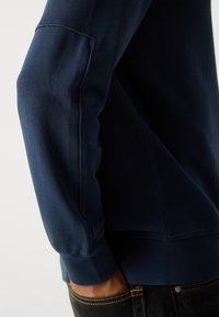 Nærbilde av en person som har på seg en mørkeblå hettegenser med synlige søm detaljer og en sidelomme, med hånden i lommen på svarte bukser.