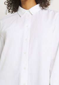 Camicia bianca con bottoni, realizzata in un tessuto liscio, caratterizzata da un colletto classico e sei bottoni sul davanti, mostrata senza strati aggiuntivi.