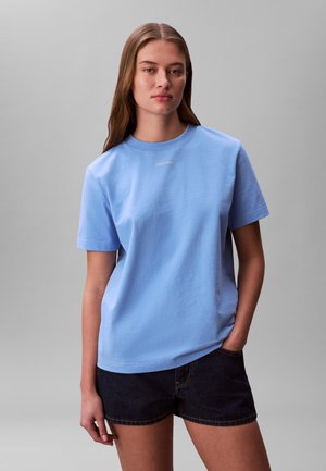 Noor naine pikkade pruunide juustega, seljas helesinine Calvin Kleini T-särk ja tumedad teksasepüksid, seisab ühetaolise halli tausta ees.