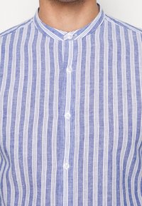 Koszula na guziki z pionowymi, niebiesko-białymi paskami, kołnierzykiem typu mandarynka i sześcioma guzikami z przodu. Tekstura bawełnianej tkaniny.