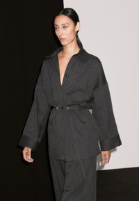 A model walks confidently in a black striped outfit with an oversized shirt and belt, set against a minimalistic backdrop.