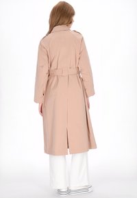 Beige trenchcoat met een ceintuur in de taille, lange mouwen en een achtersplit, gecombineerd met witte wijde broekspijpen en gestreepte sneakers.