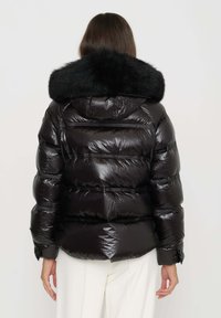 Giaccone a piumino nero con finitura lucida, dotato di cappuccio con bordo in pelliccia, trama a coste e tasche laterali. Il design è squadrato e imbottito.