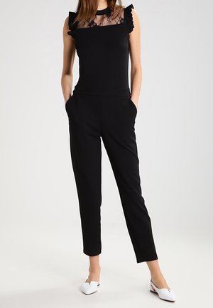 Combinaison noire avec un col en tulle froncé, haut ajusté, pantalons amples et poches latérales. Associée à des ballerines blanches à bout pointu.