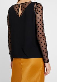 Černý top s průhlednými rukávy s puntíky, designem ve tvaru V na zádech, texturovaným tělem, doplněný o hladkou zlatou sukni. Hlavními rysy je klíčová spona.