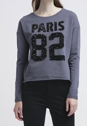 Kvinde iført grå langærmet top med sort paliettekst "PARIS 82" og sorte bukser, stående mod en ensfarvet lys baggrund.