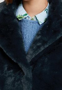 Laivastonsininen tekoturkistakki, pehmeällä ja muhkealla tekstuurilla, leveällä kauluksella ja kerroksittain vaaleansinisen neulotun villapaidan sekä kukkakauluksisen paidan päällä.