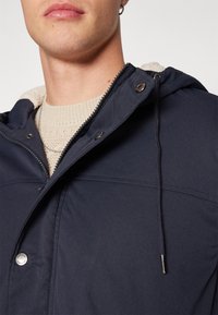 Mies pukeutuneena laivastonsiniseen hupparijakkuun, jossa on vetoketju ja painonapit, päällään beige neulepaita, kaula, huulet ja korvakoru näkyvissä.