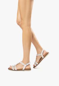 I sandali bianchi in pelle presentano dettagli con borchie, cinghie per la caviglia regolabili e una suola piatta marrone. Il design include diverse cinghie per un supporto ottimale.