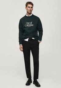Felpa verde con testo bianco "Out of Calendar" abbinata a una camicia bianca con colletto, pantaloni neri e scarpe nere. Tessuto liscio, vestibilità casual.