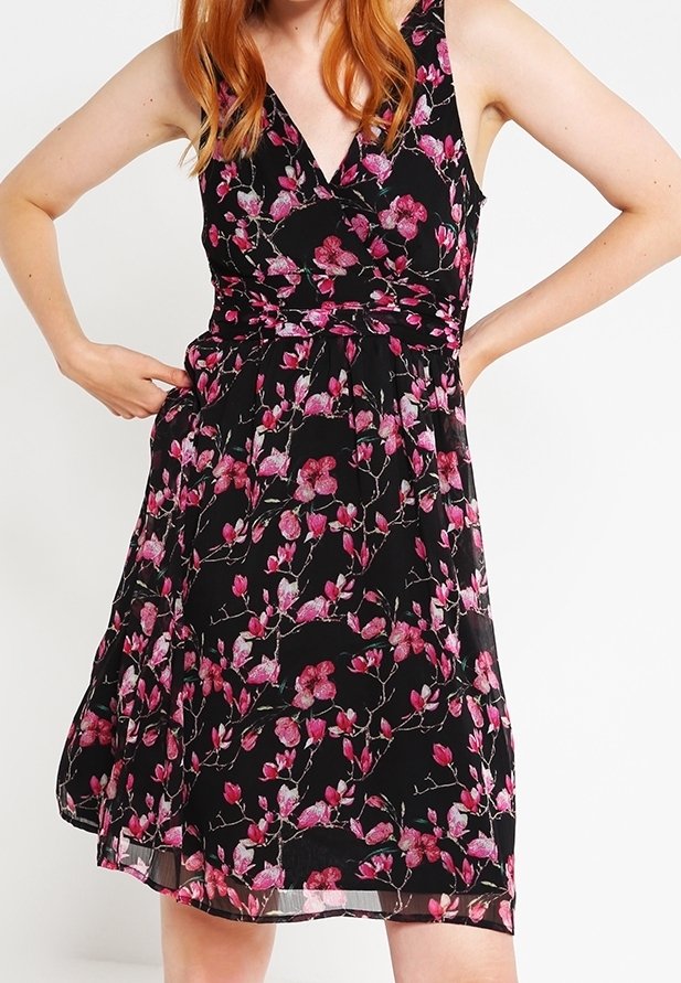Femme portant une robe noire sans manches avec un motif floral rose, mains sur les hanches, montrant les détails et la coupe de la robe.