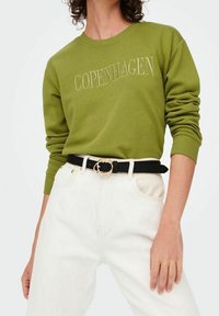 Zelená mikina s nápisem "Copenhagen" a žebrovanými manžetami, doplněná bílými džíny a černým opaskem se zlatou sponou.