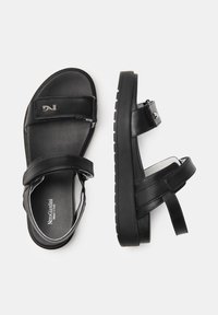 Sandali in pelle nera con due fasce larghe, cinturino regolabile, suola texturizzata e soletta con marchio, che mostrano un design minimalista.