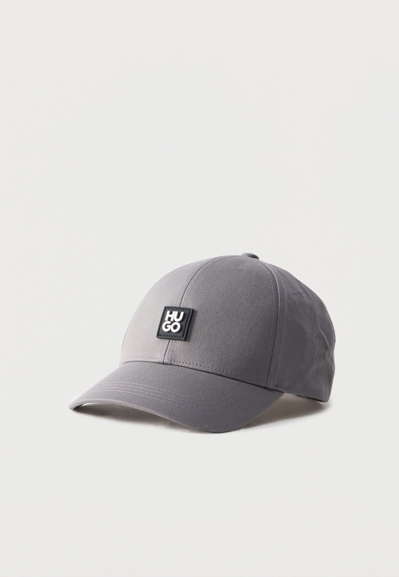 Γκρι καπέλο μπέιζμπολ με καμπυλωτό γείσο και μαύρο τετράγωνο πάτς με την ένδειξη "HUGO" στο μπροστινό μέρος, εμφανισμένο σε απλό λευκό φόντο.