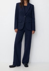 Femme portant un costume bleu marine à fines rayures avec un pantalon large et des chaussures noires à bout carré, debout devant un fond blanc.