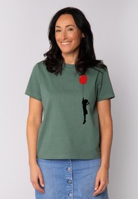 Groen katoenen t-shirt met korte mouwen, voorzien van een zwarte silhouet van een figuur die een rode ballon vasthoudt aan de voorkant.