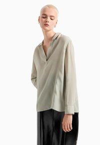 Blusa verde chiaro in tessuto setoso, con scollatura a V, maniche lunghe e orlo asimmetrico, abbinata a una gonna nera lucida.