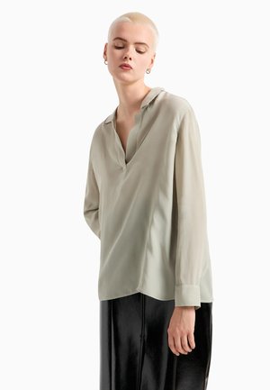Blusa verde chiaro in tessuto setoso, con scollatura a V, maniche lunghe e orlo asimmetrico, abbinata a una gonna nera lucida.