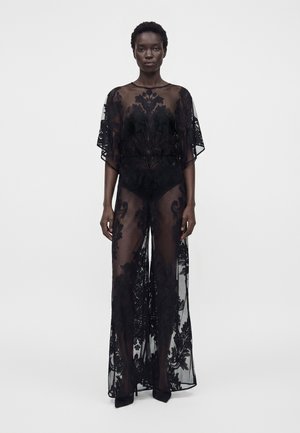 Femme portant une longue robe noire transparente avec des motifs floraux sur des sous-vêtements noirs, debout devant un fond blanc uni.