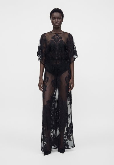 Mujer con un vestido largo negro transparente con estampados florales sobre ropa interior negra, de pie frente a un fondo blanco liso.