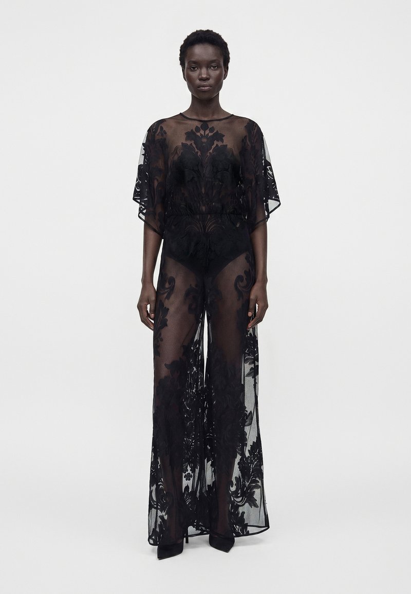 Femme portant une longue robe noire transparente avec des motifs floraux sur des sous-vêtements noirs, debout devant un fond blanc uni.