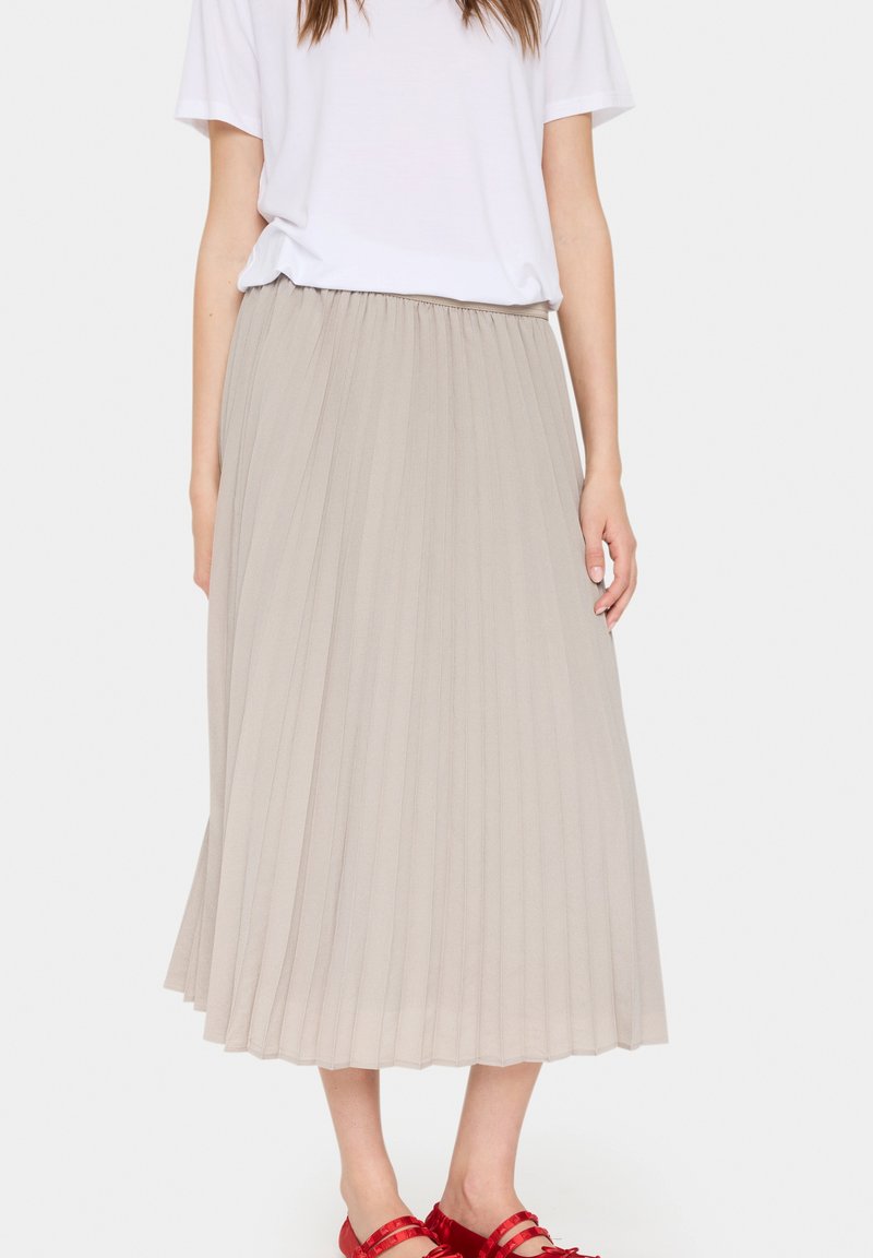 Gonna beige a vita alta, plissettata e lunga fino al ginocchio, realizzata in tessuto leggero, con una cintura elastica e abbinata a sandali rossi.