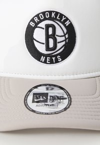 Fehér sapka fekete Brooklyn Nets logóval, amelyen egy kosárlabda és a "B" betű található. Állítható pánt, sima szürke karimával és varrási részletekkel.