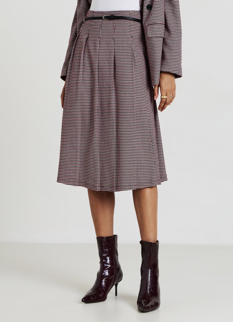 Une jupe plissée mi-longue à motif pied-de-poule avec des couleurs rouge, blanc et noir, associée à des bottines vernies bordeaux.