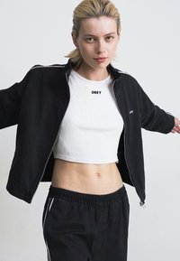 Ungdom med blondt hår iført en svart jakke med glidelås over en hvit cropped topp og svarte joggebukser, som står med armene utstrakt.