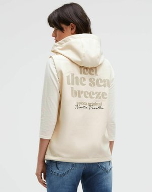 Frau trägt eine cremefarbene ärmellose Kapuzenweste mit dem Text "feel the sea breeze" auf dem Rücken über einem weißen Dreiviertelarm-Shirt und blauen Jeans.