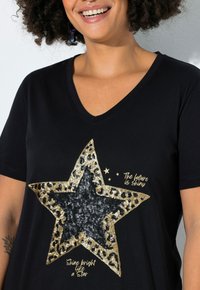 Personne souriante portant un t-shirt noir à col en V avec une étoile en sequins et un texte doré indiquant "L'avenir est brillant" et "Brille comme une étoile."