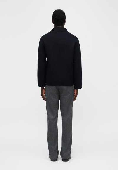 Άνδρας που στέκεται με την πλάτη προς τα εμπρός, φοράει μαύρο μπουφάν, γκρι παντελόνι με σχέδιο και μαύρα παπούτσια, σε απλό λευκό φόντο.