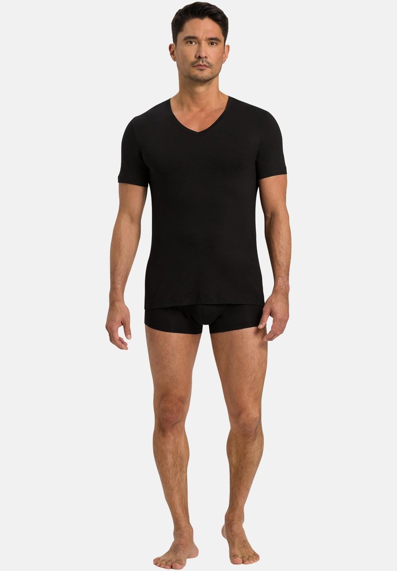 Mann steht barfuß und trägt ein schwarzes V-Ausschnitt-T-Shirt mit kurzen Ärmeln sowie schwarze, eng anliegende Boxershorts vor neutralem Hintergrund.