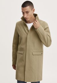 Cappotto di lana beige con colletto alto, due tasche frontali e vestibilità su misura, caratterizzato da una texture liscia e dettagli di design minimalisti.